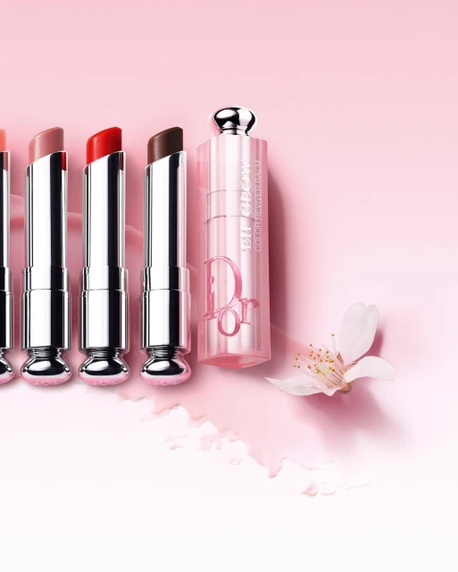 Dior lipstick collection in multiple colors displayed with a single blossom against a pink background gradient for beauty advertising.