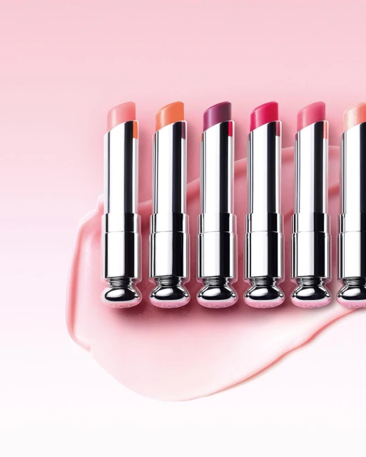 Close-up of six lipsticks in a row with varying shades of pink and purple, showcasing cosmetic beauty products against a pink background.