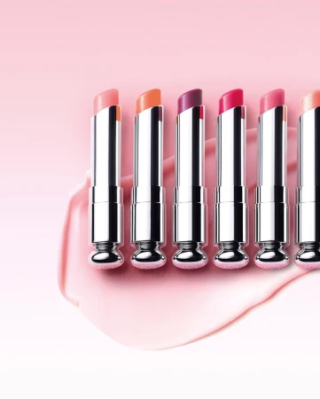 Close-up of six lipsticks in a row with varying shades of pink and purple, showcasing cosmetic beauty products against a pink background.