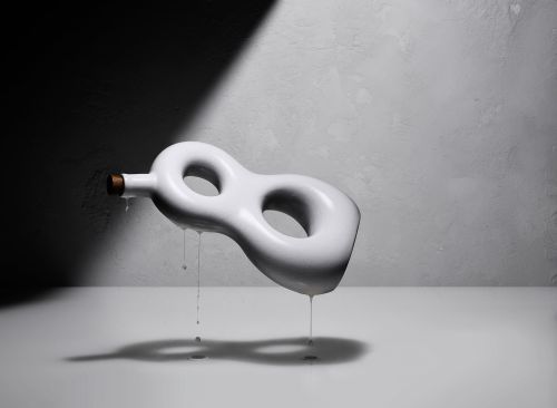 Still life shot of a white, abstract vase with liquid dripping, set against a textured gray wall with strong shadow play.