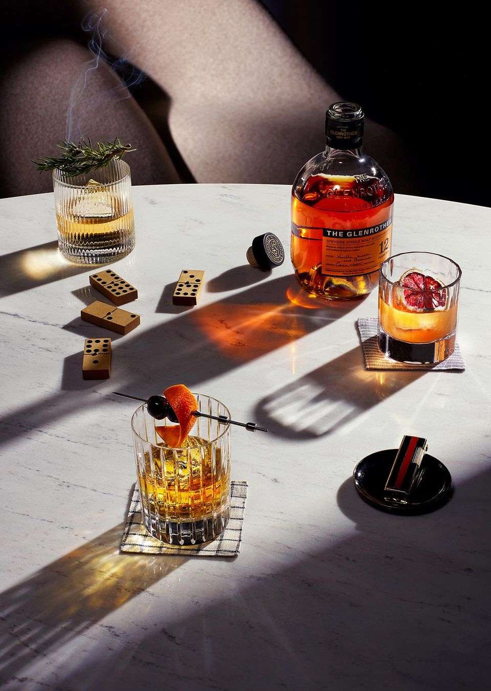 Cocktail photography on marble table featuring whiskey drinks, dominoes, garnish, and a bottle of Glenturret Scotch whiskey.
