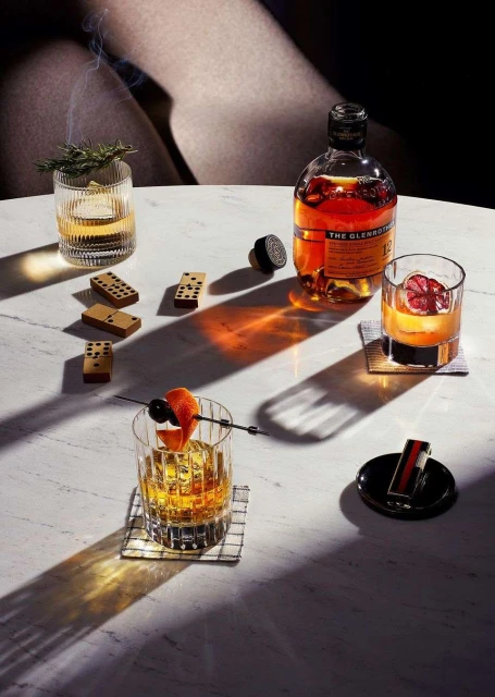Cocktail photography on marble table featuring whiskey drinks, dominoes, garnish, and a bottle of Glenturret Scotch whiskey.
