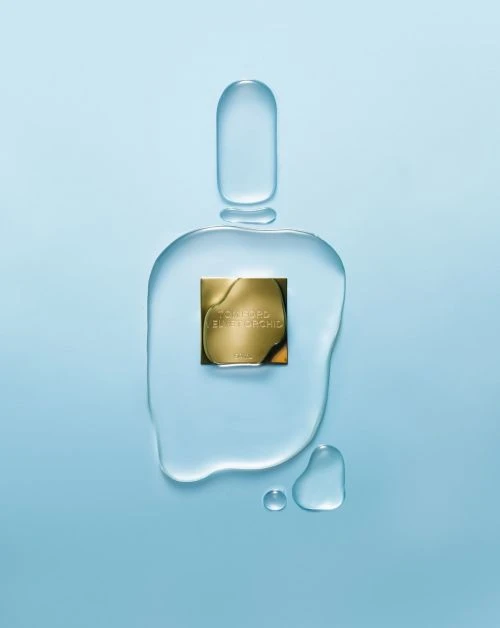 Square gold-tone perfume bottle surrounded by transparent water-like gel drops against a light blue background. Minimalist beauty shot.