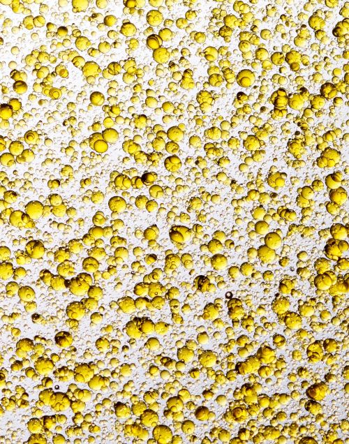 Close-up of golden yellow liquid with bubbles, resembling oil droplets. The composition features a textured pattern on a white background.