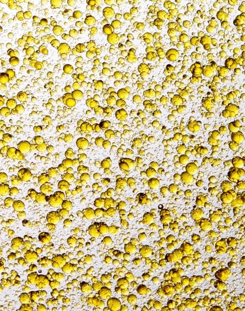 Close-up of golden yellow liquid with bubbles, resembling oil droplets. The composition features a textured pattern on a white background.