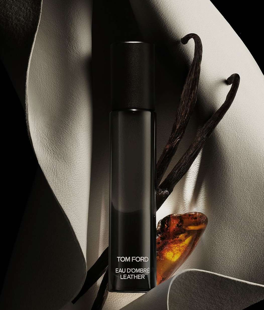 Tom Ford Eau D'Ombre Leather perfume bottle displayed with vanilla beans and amber stone on a cream-colored leather backdrop.