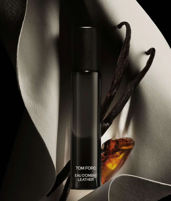 Tom Ford Eau D'Ombre Leather perfume bottle displayed with vanilla beans and amber stone on a cream-colored leather backdrop.