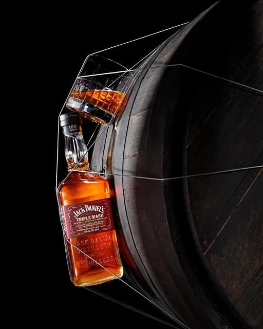 Bottle of Jack Daniel's Triple Mash Whiskey balanced on a barrel with a glass in a minimalist wire frame against a black backdrop.