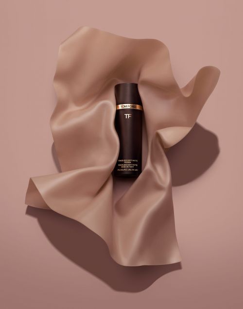 A bottle of Tom Ford beauty product is placed on a light peach silky fabric against a matching peach background in a product shot.