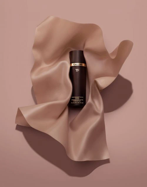 A bottle of Tom Ford beauty product is placed on a light peach silky fabric against a matching peach background in a product shot.