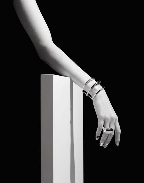 Close-up black and white shot of a woman's arm resting on a white cube, adorned with silver bangles and a statement ring against a black background.