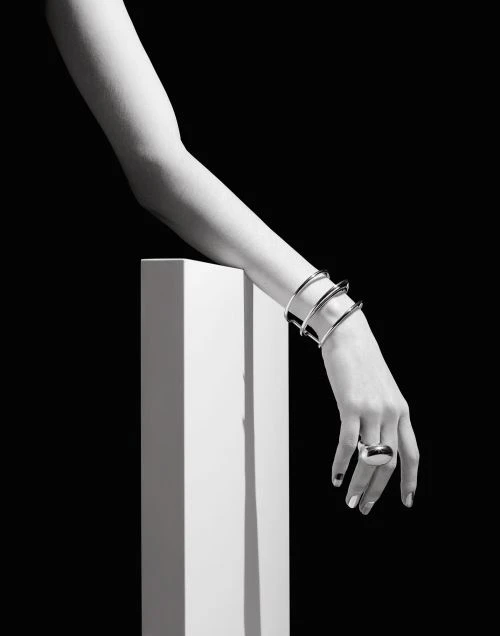 Close-up black and white shot of a woman's arm resting on a white cube, adorned with silver bangles and a statement ring against a black background.