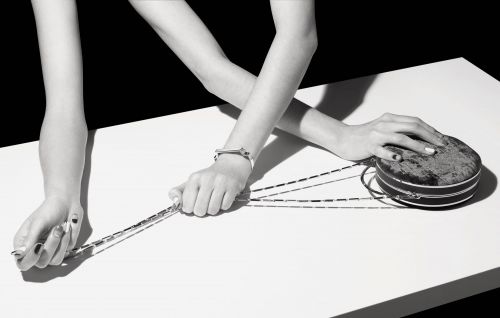 Monochrome shot of a round clutch with chain handle, resting on a white surface, held by an adult woman's hands with painted nails.