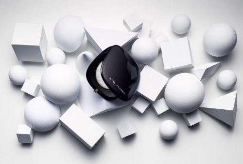 High-angle shot of a black Marc Jacobs compact surrounded by white geometric shapes, including spheres, cubes, and triangles on a white surface.