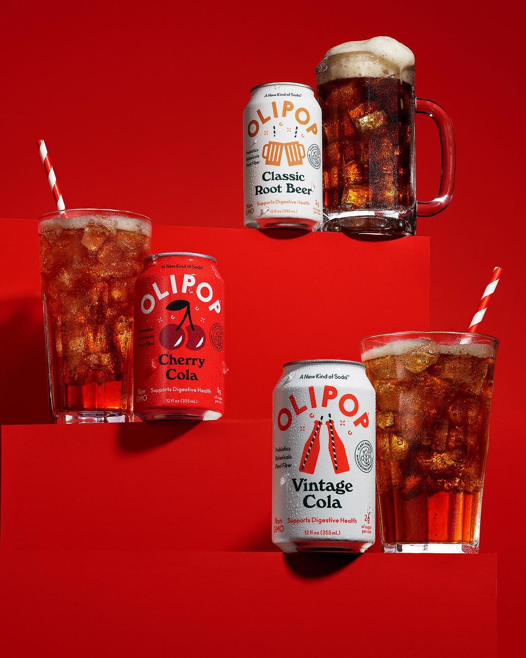 Three varieties of Olipop soda cans alongside matching glass mugs and glasses filled with the dark soda, on a red background.