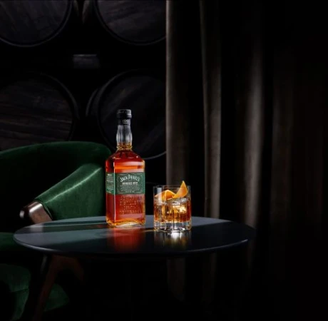 Jack Daniel's whiskey bottle and glass with orange peel garnish on a black table, barrels and dark curtain in background. 