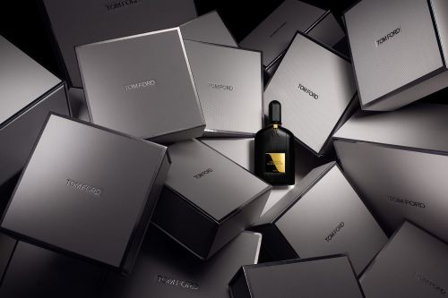 A black perfume bottle surrounded by a chaotic stack of grey branded boxes under dramatic studio lighting.