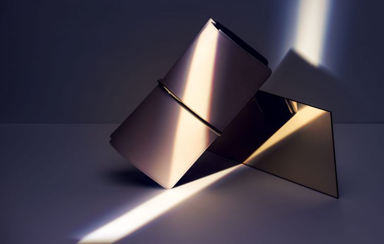 A beige rectangular clutch purse on a golden metal frame casting shadows on a light grey background in a minimalist setting.