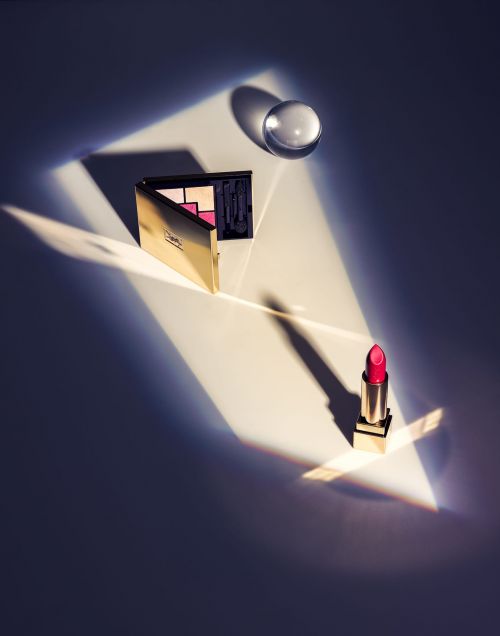Beauty still life: eyeshadow palette, red lipstick tube, and glass orb. Cosmetics arranged in beam of light on a white background.
