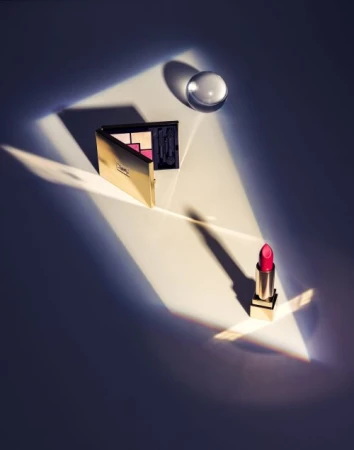 Beauty still life: eyeshadow palette, red lipstick tube, and glass orb. Cosmetics arranged in beam of light on a white background.