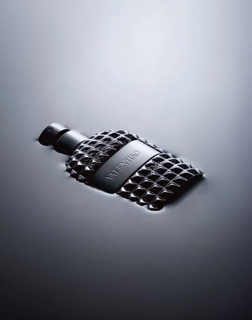 A bottle of Valentino perfume with a studded design, partially submerged in water, is prominently displayed in a sleek, monochromatic composition.