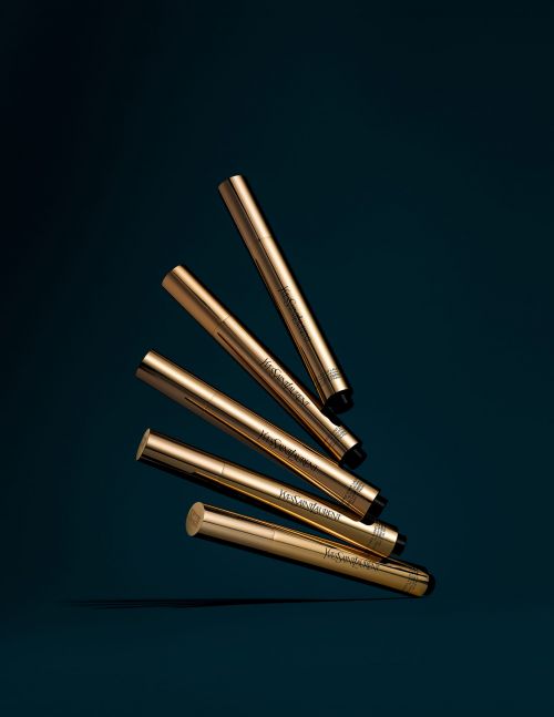 Group of golden YSL Touche Éclat illuminating pens displayed on a dark teal background in a beauty product advertisement.