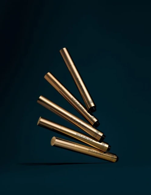 Group of golden YSL Touche Éclat illuminating pens displayed on a dark teal background in a beauty product advertisement.