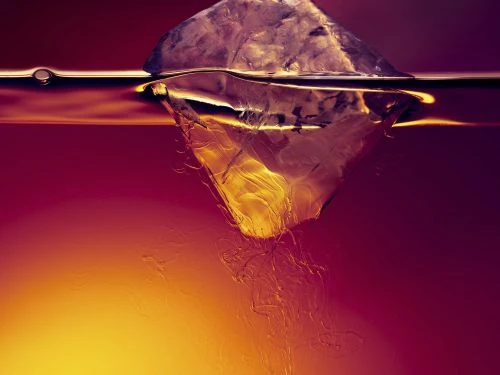 Close-up shot of an ice cube melting into a maroon liquid, lit from beneath, creating a warm glow and visible textural details.