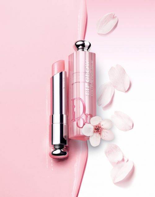 Close-up product shot of a pink Dior Addict lipstick with silver accents, surrounded by delicate white flower petals against a soft pink and white gradient backdrop.