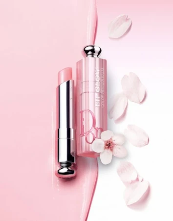 Close-up product shot of a pink Dior Addict lipstick with silver accents, surrounded by delicate white flower petals against a soft pink and white gradient backdrop.