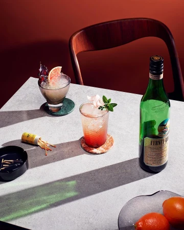Two cocktails with ice, bottle, citrus fruit, matches, and ash tray on a table, indoor setting with brown background.