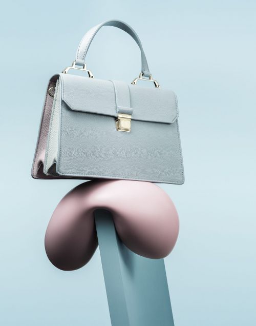 Pale blue textured leather satchel handbag with gold hardware and handle, balanced on a pink sculptural object against a blue background.