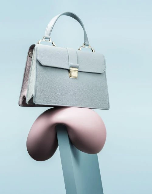 Pale blue textured leather satchel handbag with gold hardware and handle, balanced on a pink sculptural object against a blue background.