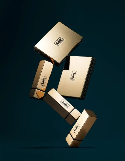 Gold Yves Saint Laurent makeup products including compacts and lipsticks floating in a dark teal studio environment.