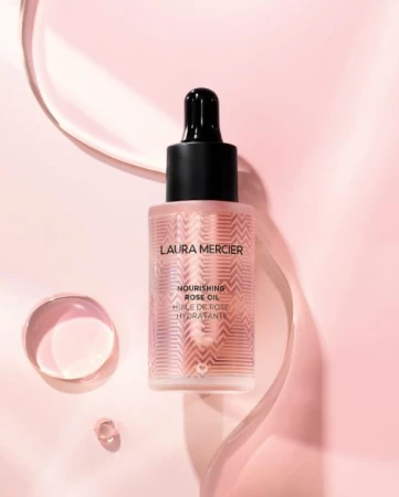 Product shot of Laura Mercier Nourishing Rose Oil in a glass bottle with a black dropper, displayed on a pink background with clear water elements.