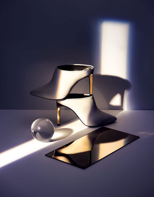 Elegant white heeled mules are stacked atop each other next to a reflective gold plate and a clear glass sphere, illuminated by light.