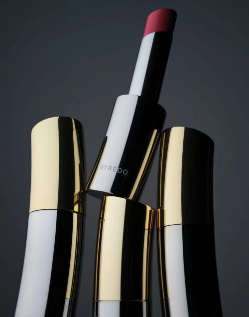 Product shot of Byredo lipstick tubes featuring gold, black, and white design with one extended tube revealing the pink lipstick.