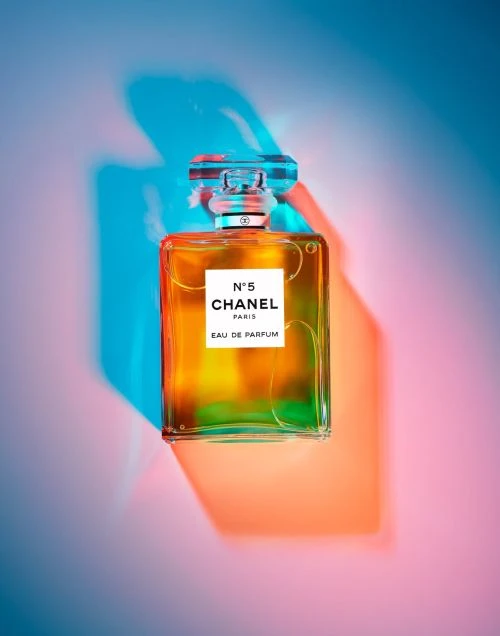 Chanel No. 5 perfume bottle featuring an amber liquid with clear glass stopper, set against a backdrop of teal and orange gradients.
