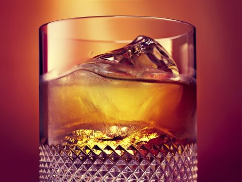 Close-up of a cut crystal glass filled with amber-colored liquor and ice, set against a warm, blurred orange background.