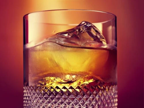 Close-up of a cut crystal glass filled with amber-colored liquor and ice, set against a warm, blurred orange background.