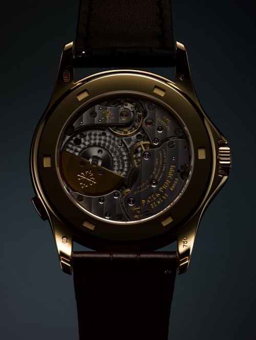 Close-up of the back of a gold wristwatch, displaying its intricate inner workings, gears, and a brown leather strap on a dark backdrop.