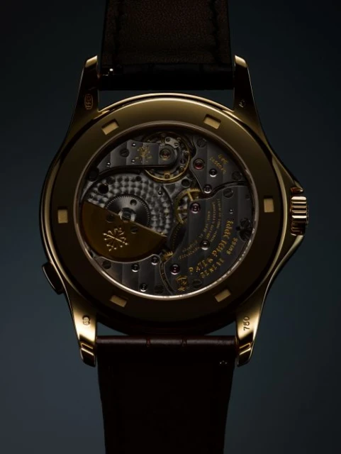 Close-up of the back of a gold wristwatch, displaying its intricate inner workings, gears, and a brown leather strap on a dark backdrop.