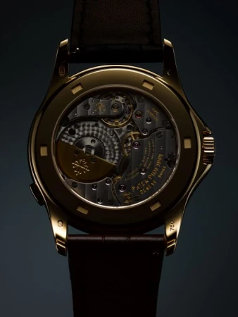 Close-up of the back of a gold wristwatch, displaying its intricate inner workings, gears, and a brown leather strap on a dark backdrop.