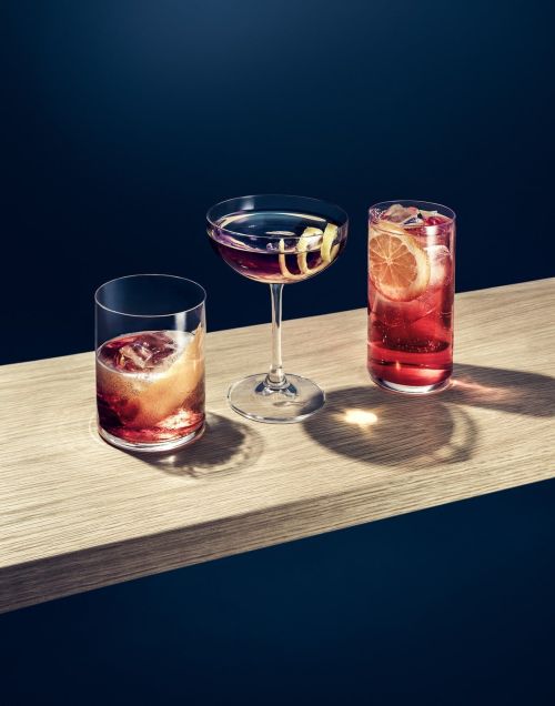 Three cocktails in clear glasses sit on a light wood surface against a dark blue background, showcasing a variety of drink presentations.