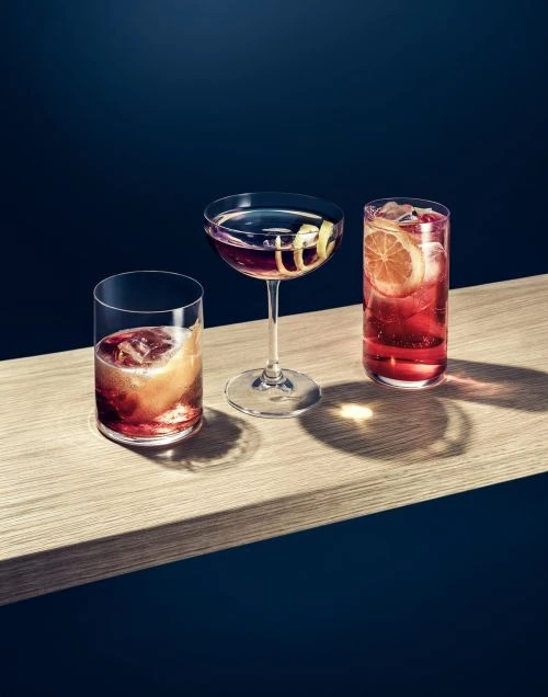 Three cocktails in clear glasses sit on a light wood surface against a dark blue background, showcasing a variety of drink presentations.
