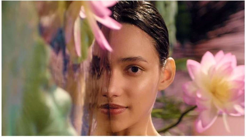 Close-up of an adult woman's face with fair skin, glossy dark hair, and neutral expression, set against a backdrop of pink lotus flowers.