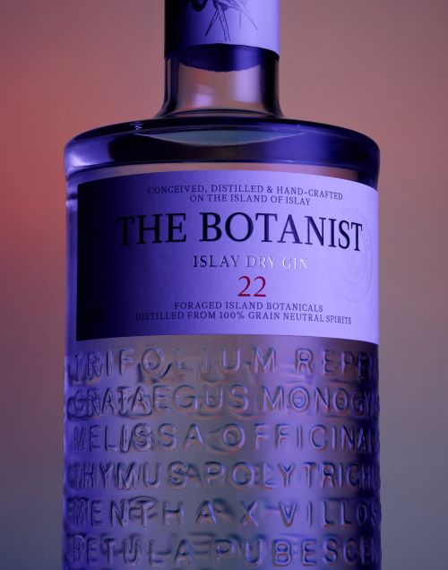Close-up studio shot of The Botanist Islay Dry Gin bottle with botanical name engravings against an orange and blue gradient background.