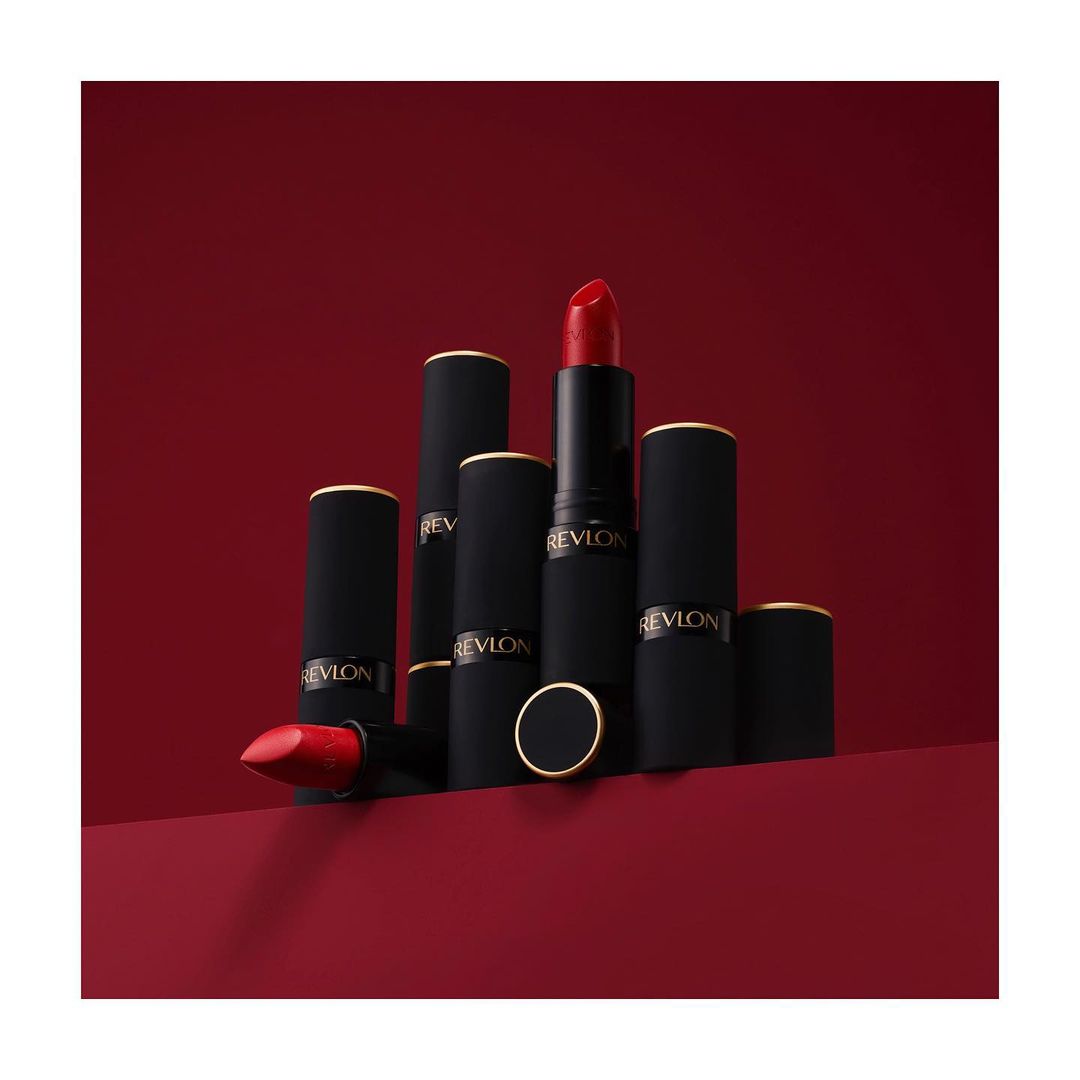 Revlon lipstick products in black and gold tubes with red lipstick, presented on a matching red surface against a red backdrop.