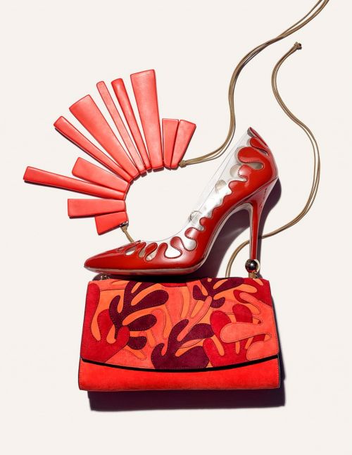 Red high-heel shoe with unique fan-like accessory and patterned handbag, presented in a studio setting against a neutral background.