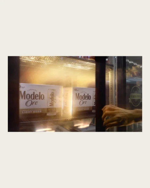 Two white 12-packs of Modelo Oro light beer are visible in a refrigerated case, with a person's hand reaching toward them.
